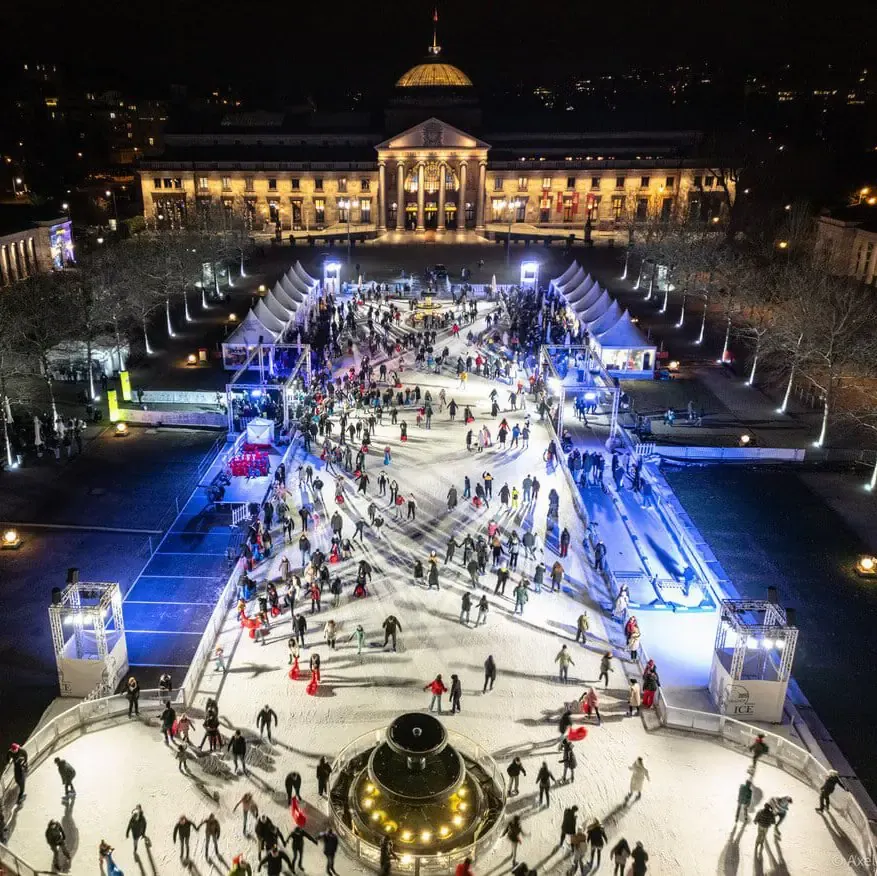 Luftaufnahme der Eisbahn Wiesbaden on Ice vor dem Kurhaus bei Nacht, voller Schlittschuhläufer unter Lichterketten. – Wiesbaden on Ice