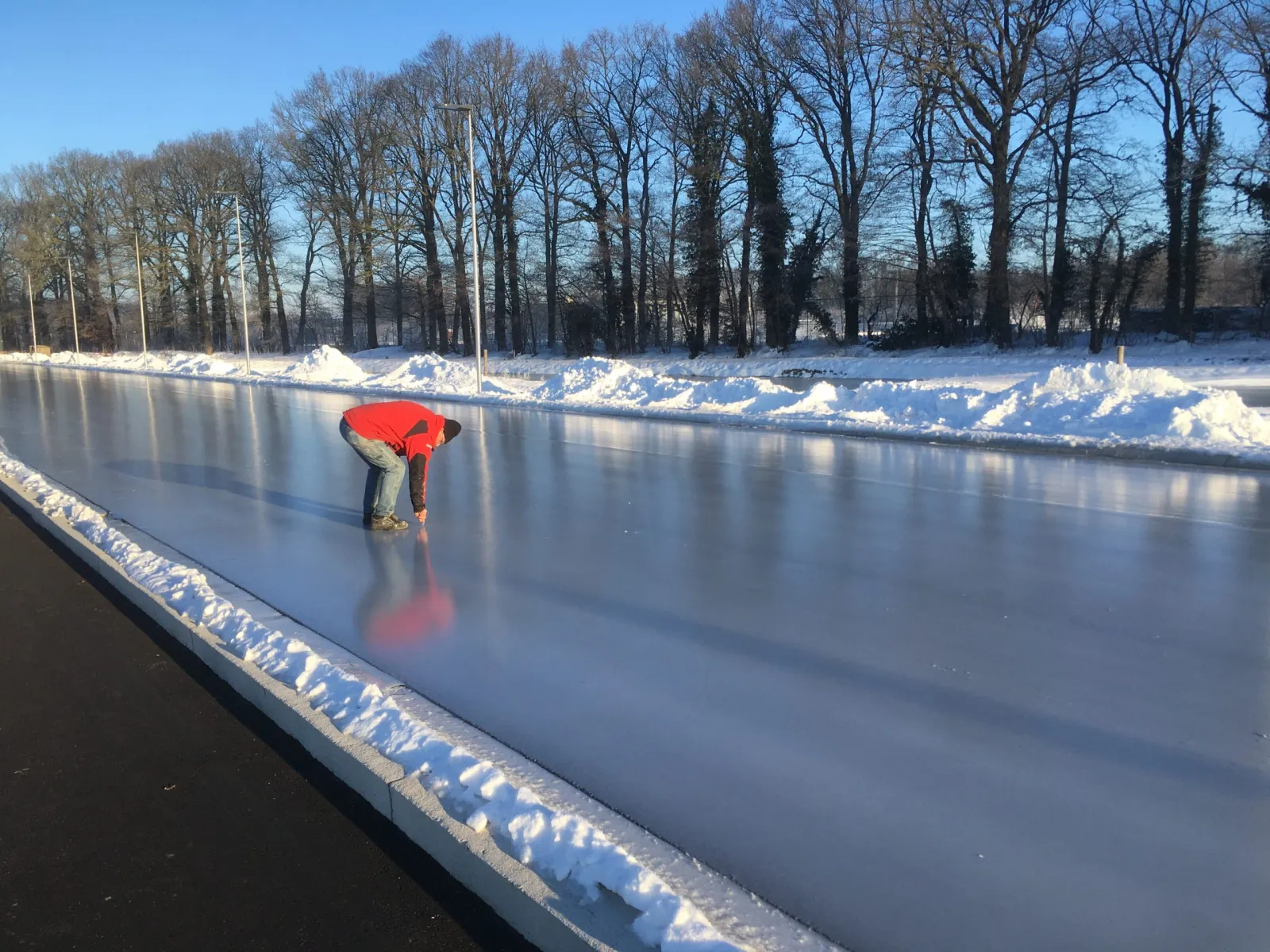 Glad zwart ijs op de 400 meter lange sproeiijsbaan van De Witte Poorte op een heldere winterochtend, met een schaatser die zich aan de rand buigt, sneeuwwallen en een rij kale bomen op de achtergrond. – WIJV De Witte Poorte