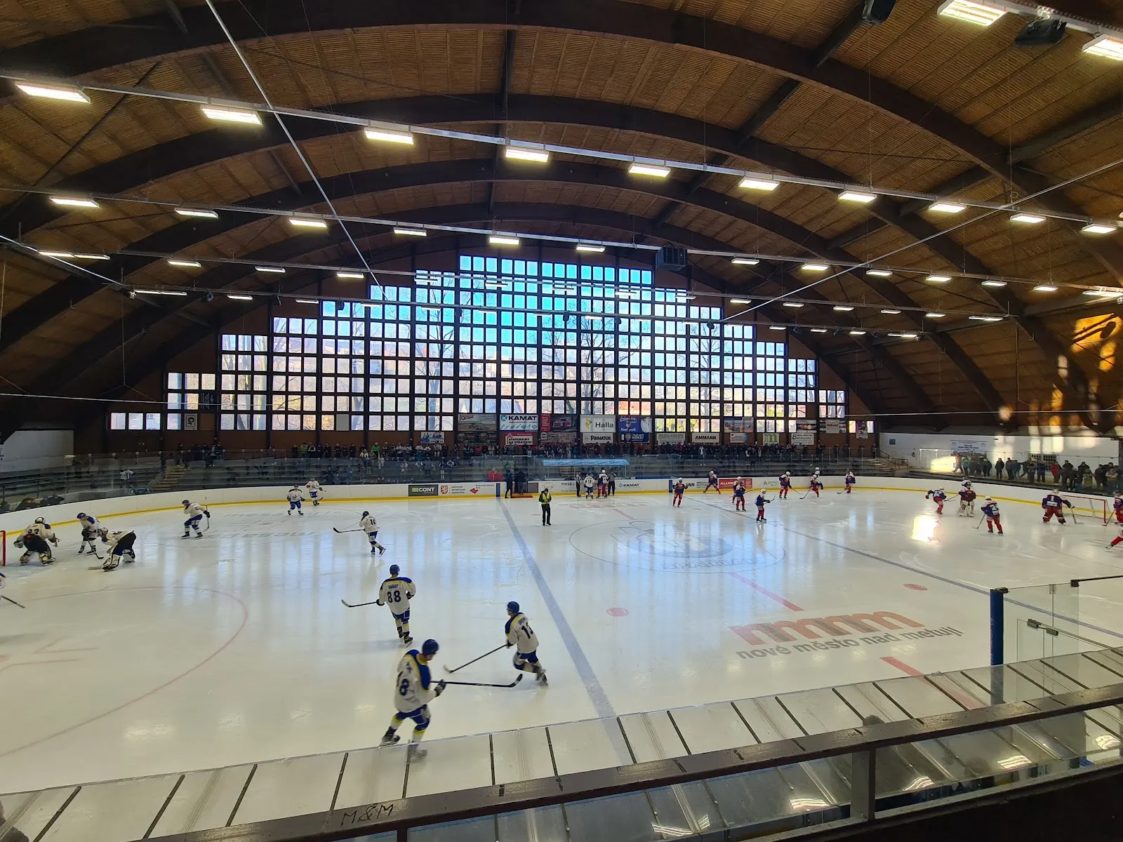 The ice sheet of the Wikov Aréna in Hronov from the stands, under the curved timber roof, with a game in progress far across the ice — Zimní stadion Hronov (Wikov aréna)
