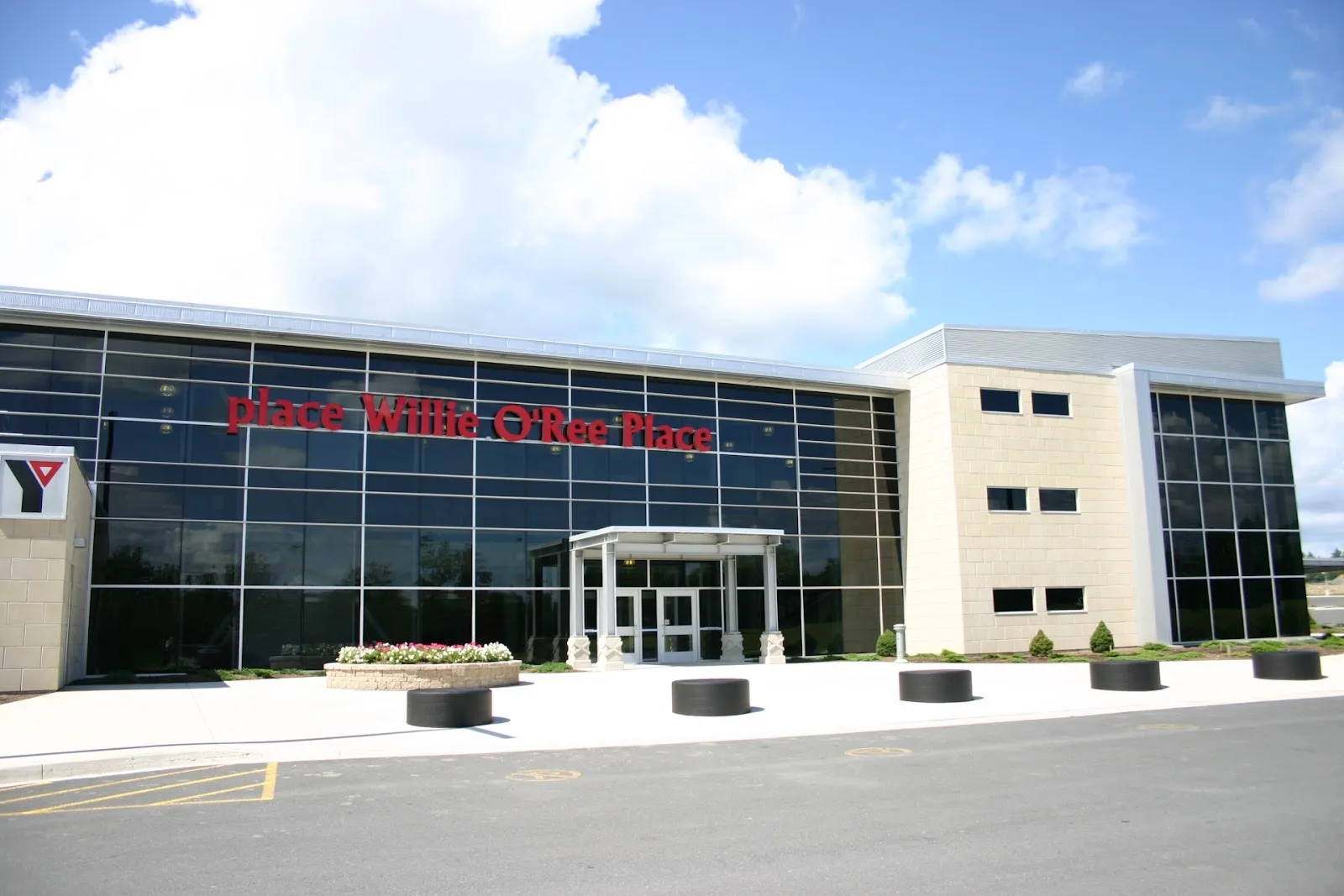 Willie O'Ree Place, a tall glass front with its name in red letters, a covered entrance and puck-shaped bollards along the walk — Willie O'Ree Place