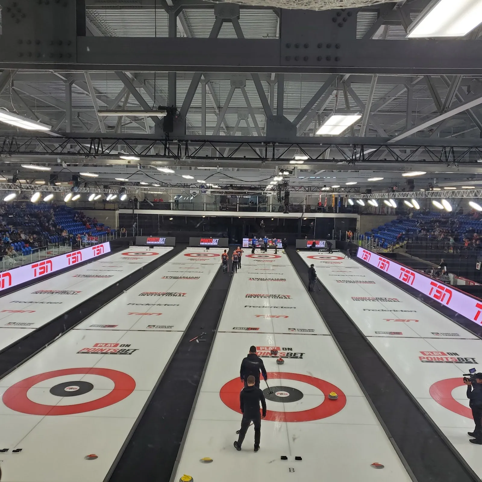 An arena bowl with a curling session across the ice, long lanes and red rings under a steel roof with purple lights along the boards. — Willie O'Ree Place