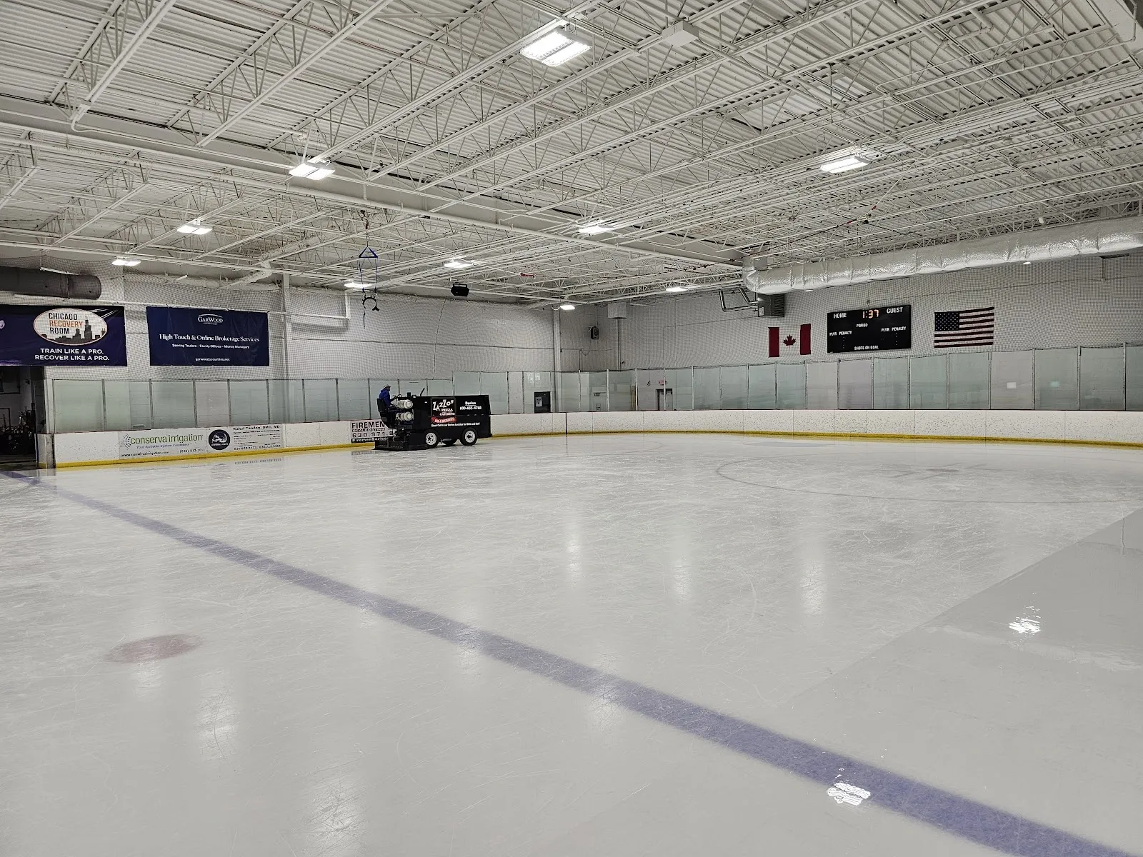 Una pista amplia y vacía vista desde las vallas, con una línea azul sobre el hielo y banderines del club y banderas en la pared del fondo, bajo un techo con estructura de acero. — Willowbrook Ice Arena