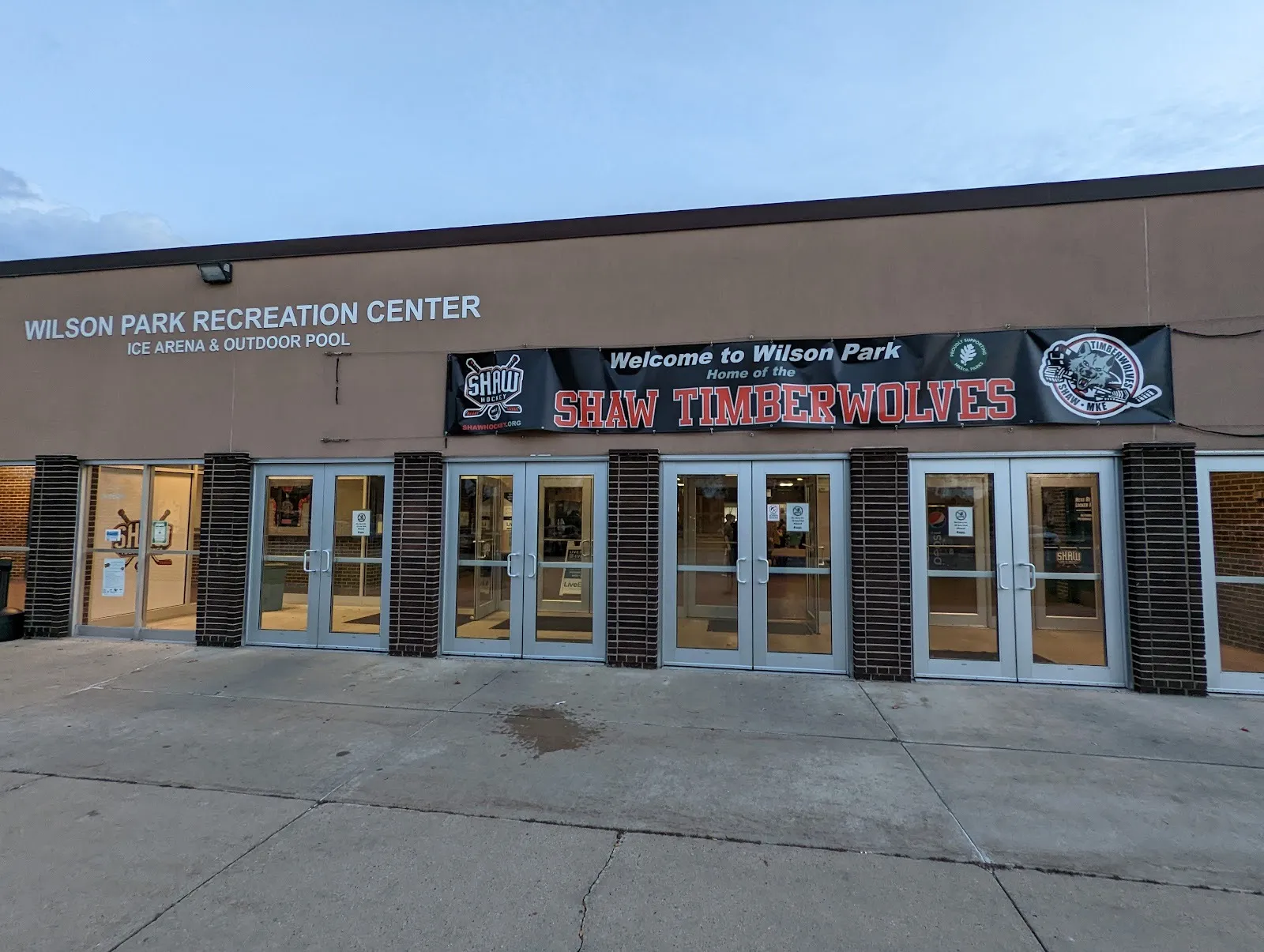 The Wilson Park Recreation Center front, Ice Arena and Outdoor Pool lettered beside a row of glass doors and a team banner — Wilson Park Ice Arena