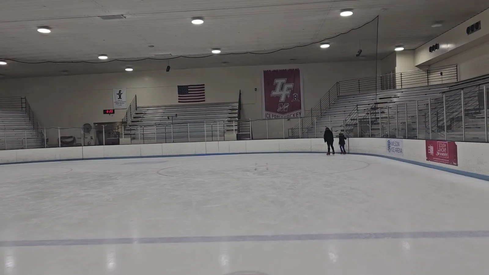The near-empty Wilson Ice Arena rink, two skaters at the far boards below grey bleachers, a flag and a hockey banner — Wilson Park Ice Arena