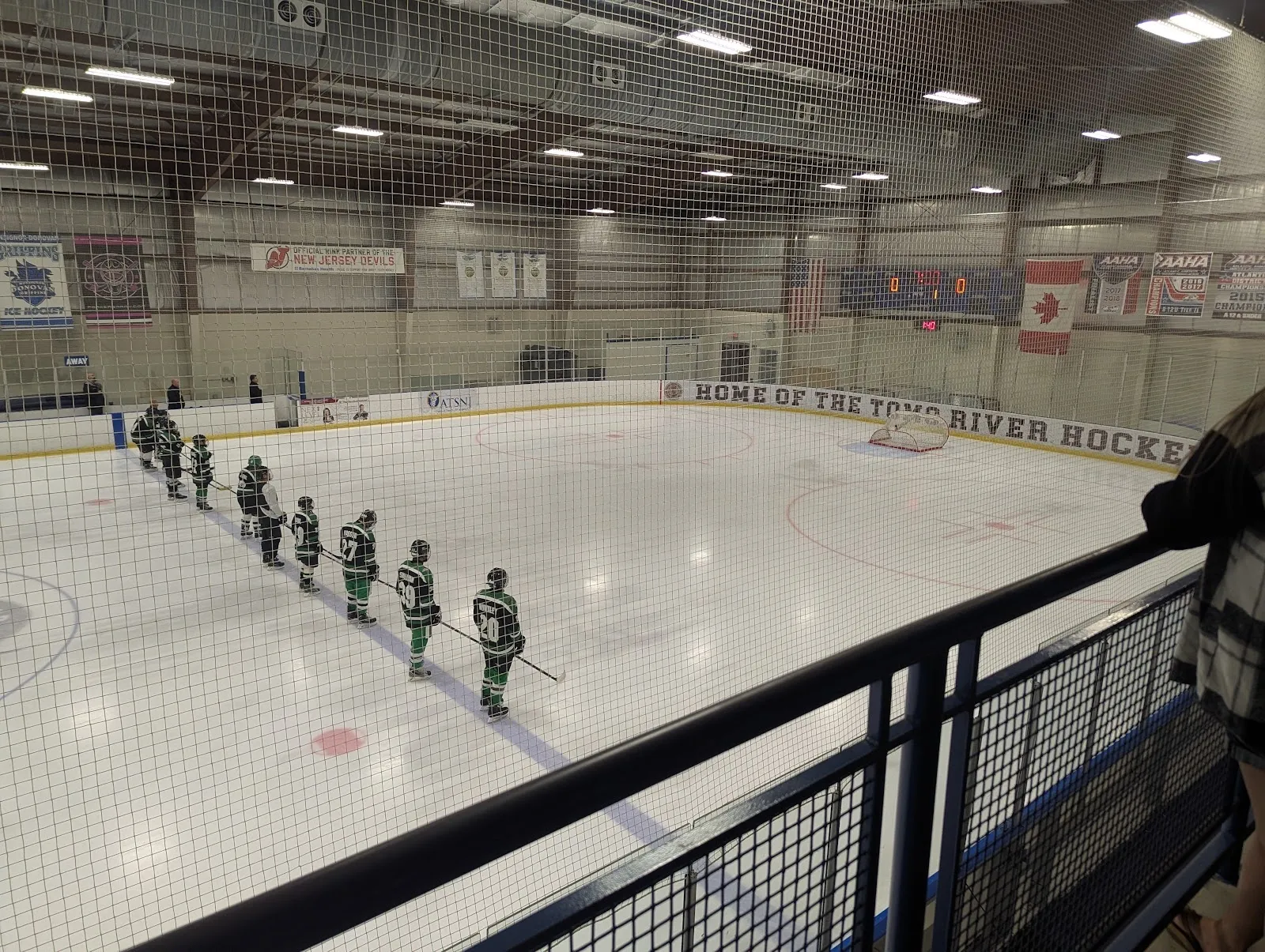 A team lined up along the blue line of a wide sheet seen over a mesh rail, banners and flags hung along the far wall under a steel roof. — Winding River Skating Center