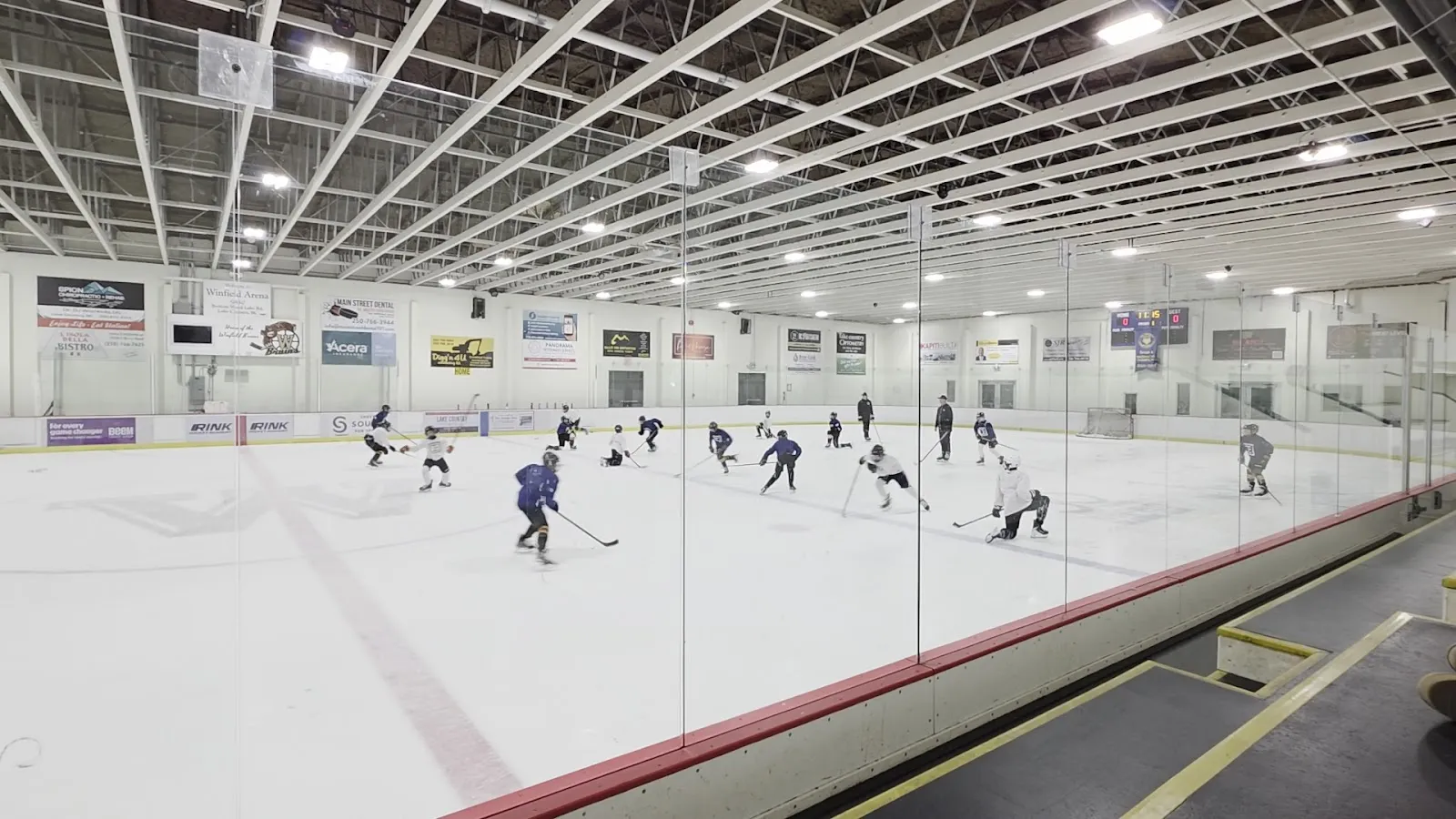 The Winfield Arena ice seen through the glass, with skaters on the ice, the white boards and the steel roof structure — Winfield Arena