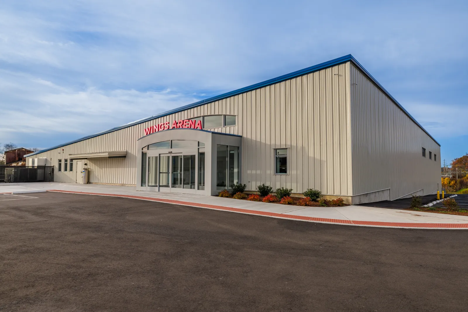 The long beige metal arena building with red "Wings Arena" lettering over the arched glass entrance, seen across the parking lot. — Wings Arena