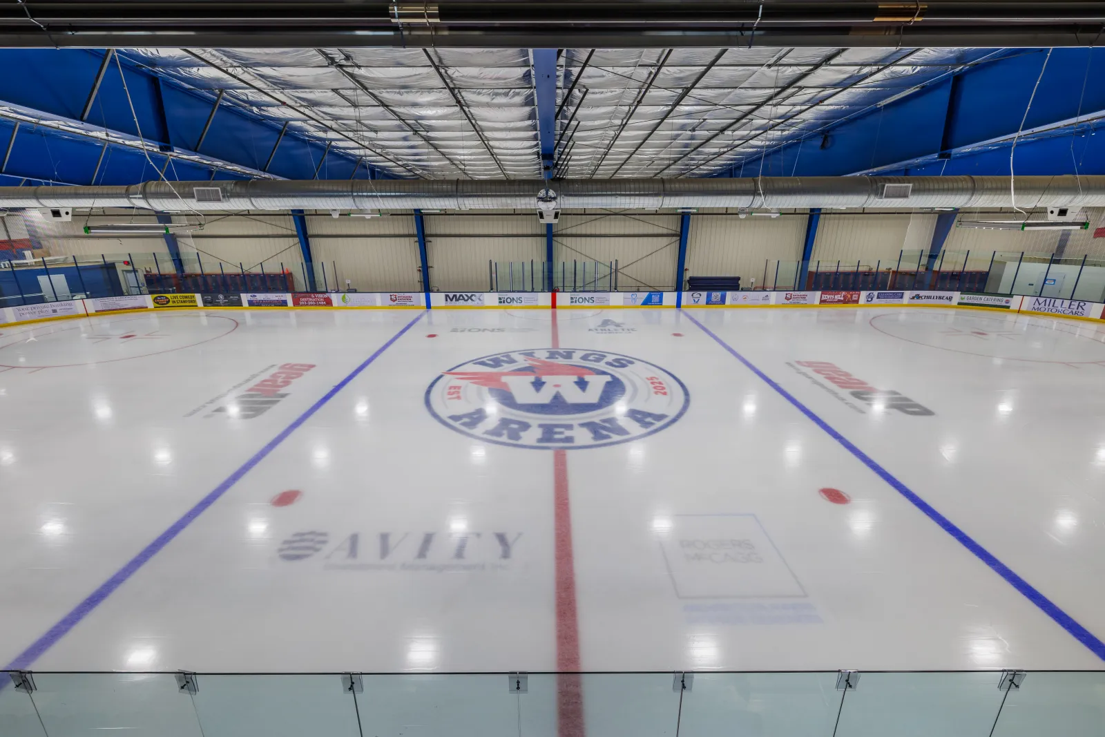 The empty rink from center ice with the Wings Arena crest at center, blue lines and a blue-trussed roof overhead. — Wings Arena