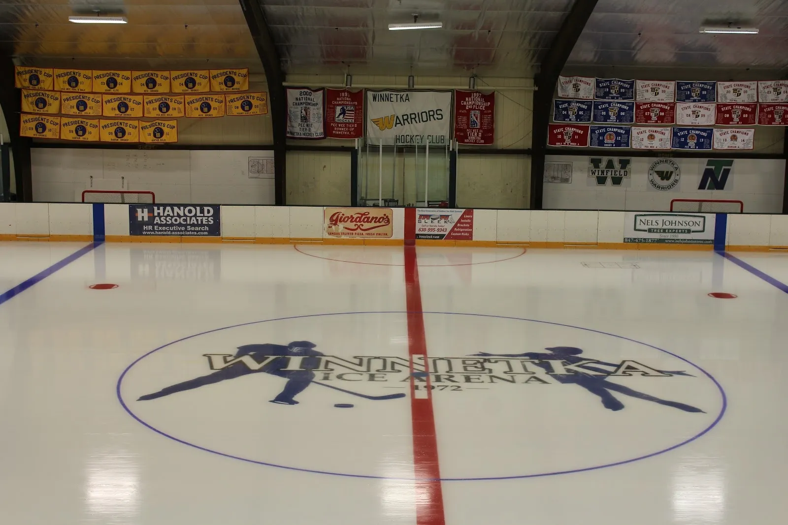 Una pista vacía con el escudo de la arena en el centro del hielo y una línea roja, con anuncios de patrocinadores y estandartes del club en la pared del fondo, detrás del vidrio. — Winnetka Ice Arena
