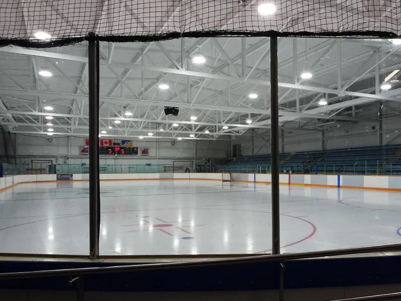 Empty indoor ice sheet seen through the players' bench glass, with overhead steel roof trusses, a scoreboard, and blue bleacher seating along the far side. — Ab McDonald Arena
