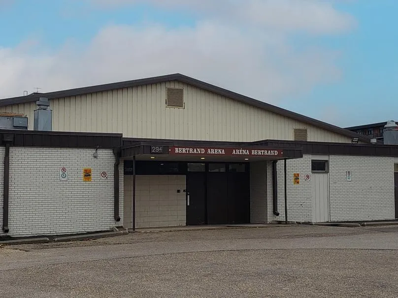 Front entrance of a low brick and metal-sided arena building, with a sign reading '294 – BERTRAND ARENA – ARÉNA BERTRAND' above the dark entry doors. — Bertrand Arena