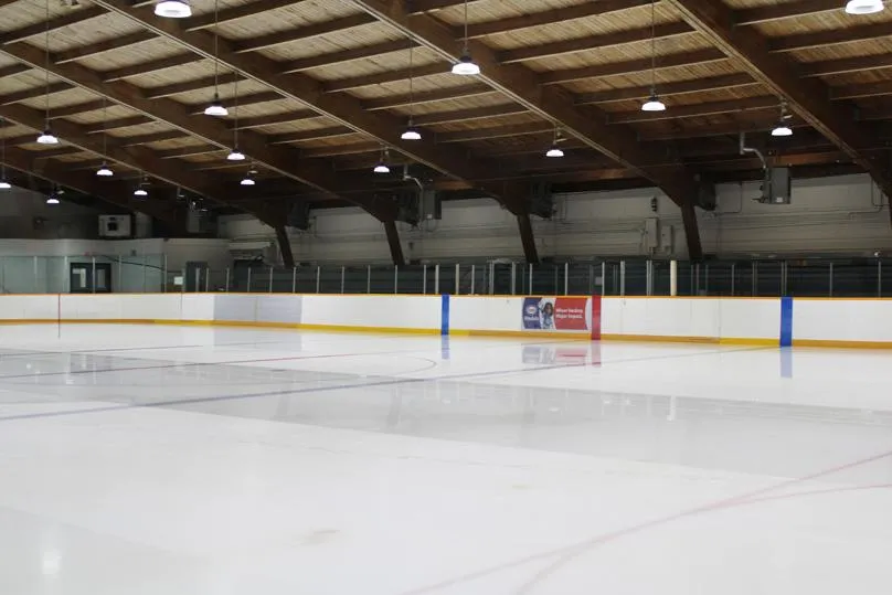 Empty indoor ice sheet under a wood-beamed roof, viewed from ice level with rink boards, a sponsor ad board, and low bleacher seating along the far wall. — Bertrand Arena