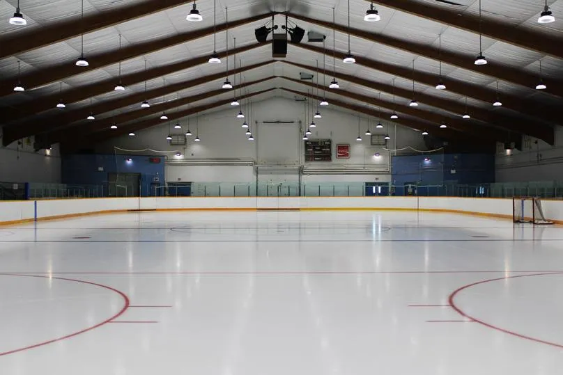 Pista de hielo techada vacía bajo un techo en forma de A de vigas de madera, vista desde el extremo de la portería con un marcador, un anuncio de Coca-Cola y porterías de hockey visibles en el hielo. — Charlie Gardiner Arena