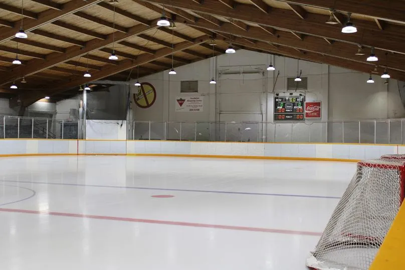 Empty indoor ice sheet under a wood-beamed roof, viewed from ice level with a scoreboard, a Coca-Cola ad board, and a goal net in the foreground. — Maginot Arena