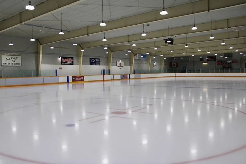 Empty ice sheet at Sam Southern Arena seen from behind the boards, with a rink-side sponsor banner and Canadian flag on the far wall. — Sam Southern Arena