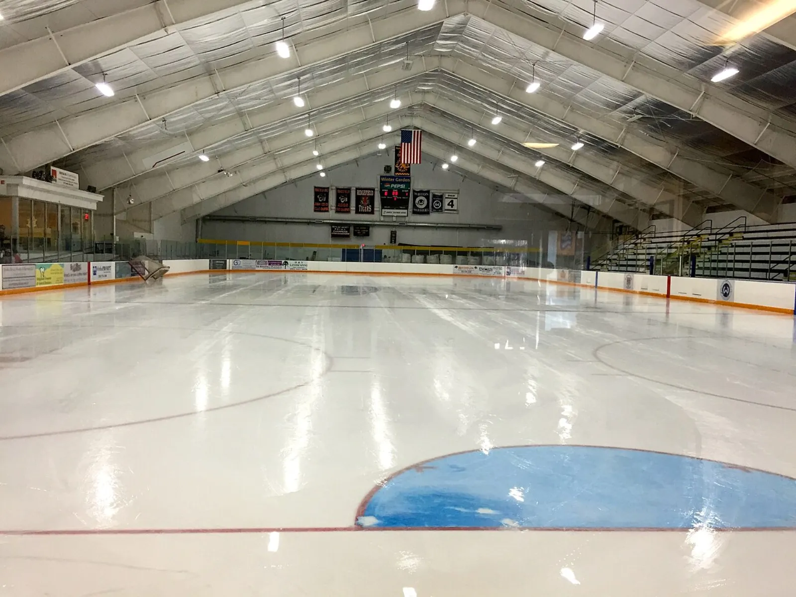 Pista de hielo vacía bajo el techo en arco de la arena, con una bandera de Estados Unidos y mantas de campeonato en el muro del fondo y 'Winter Garden' visible en el marcador. — Winter Garden Ice Arena