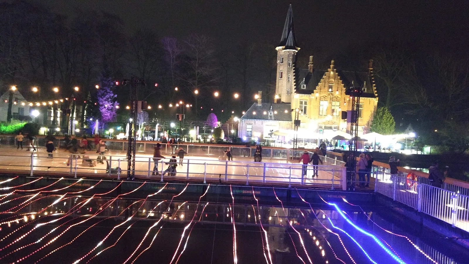 The rink at night in the Minnewater park, ringed by strings of lights, with the lit neo-Gothic castle building behind and its lights reflected in the water. — Winter Glow Ice Rink Bruges