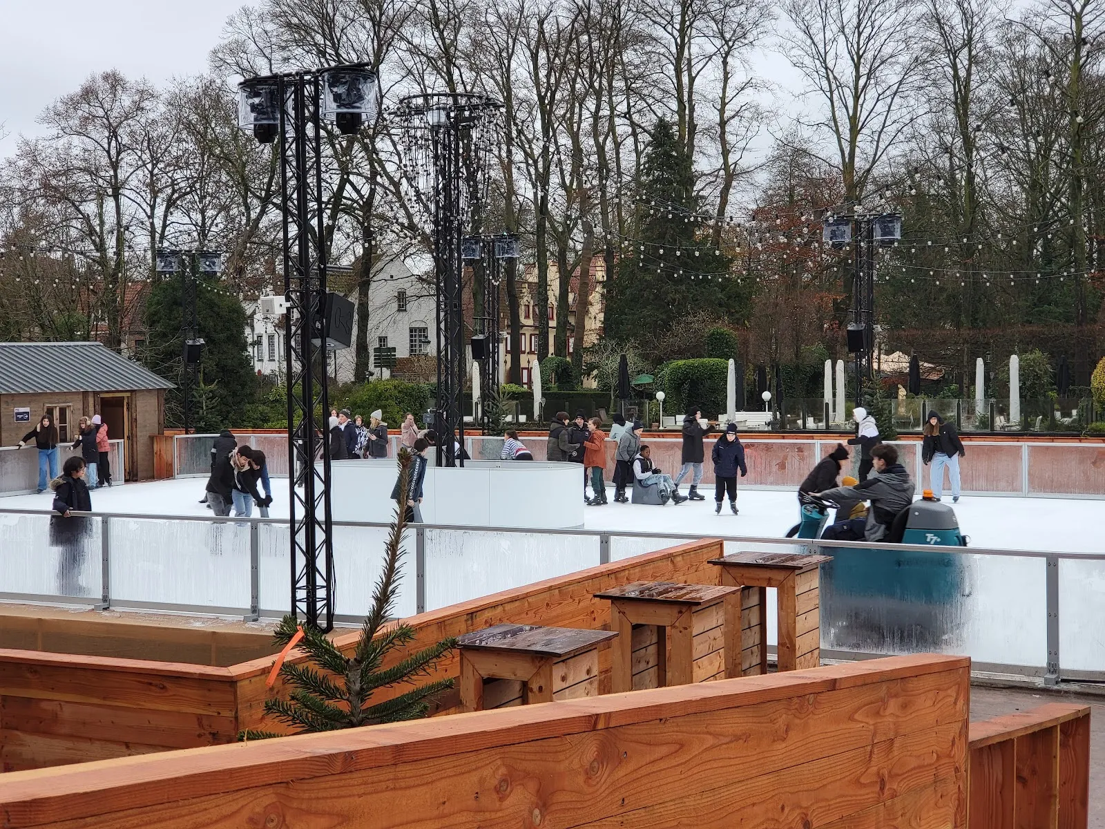 The synthetic ice rink in the park by day with skaters spread across it, light-rigging towers, a timber terrace in the foreground and bare trees behind. — Winter Glow Ice Rink Bruges