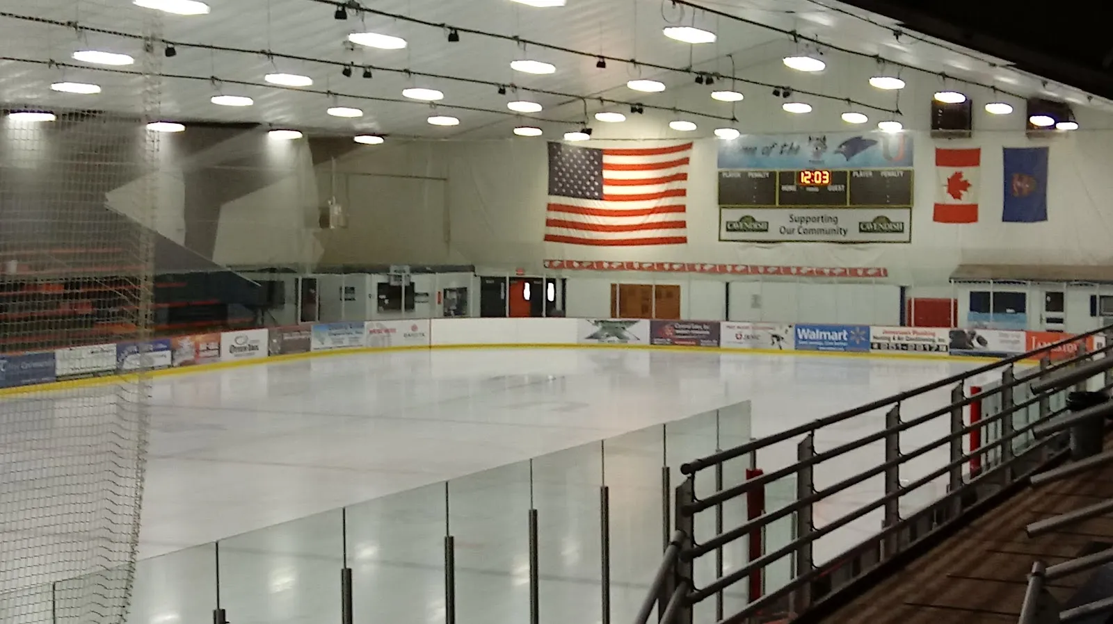 La pista vacía vista desde las gradas a través del cristal, con banderas de Estados Unidos, Canadá y del estado en la pared del fondo, un marcador y anuncios de patrocinadores a lo largo de las vallas. — Winter Sports Building