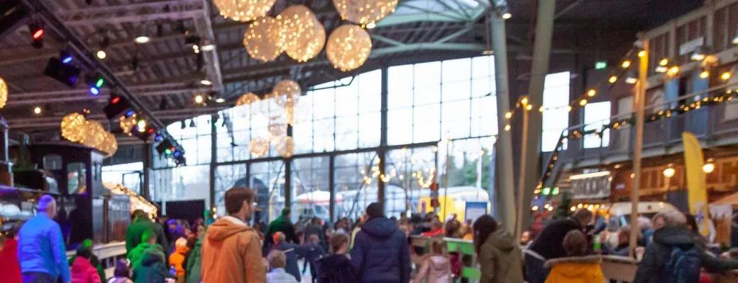 Skaters on the ice rink in the museum hall during Winter Station, with an antique steam locomotive and string lights behind them — Winter Station Ice Rink (Spoorwegmuseum)