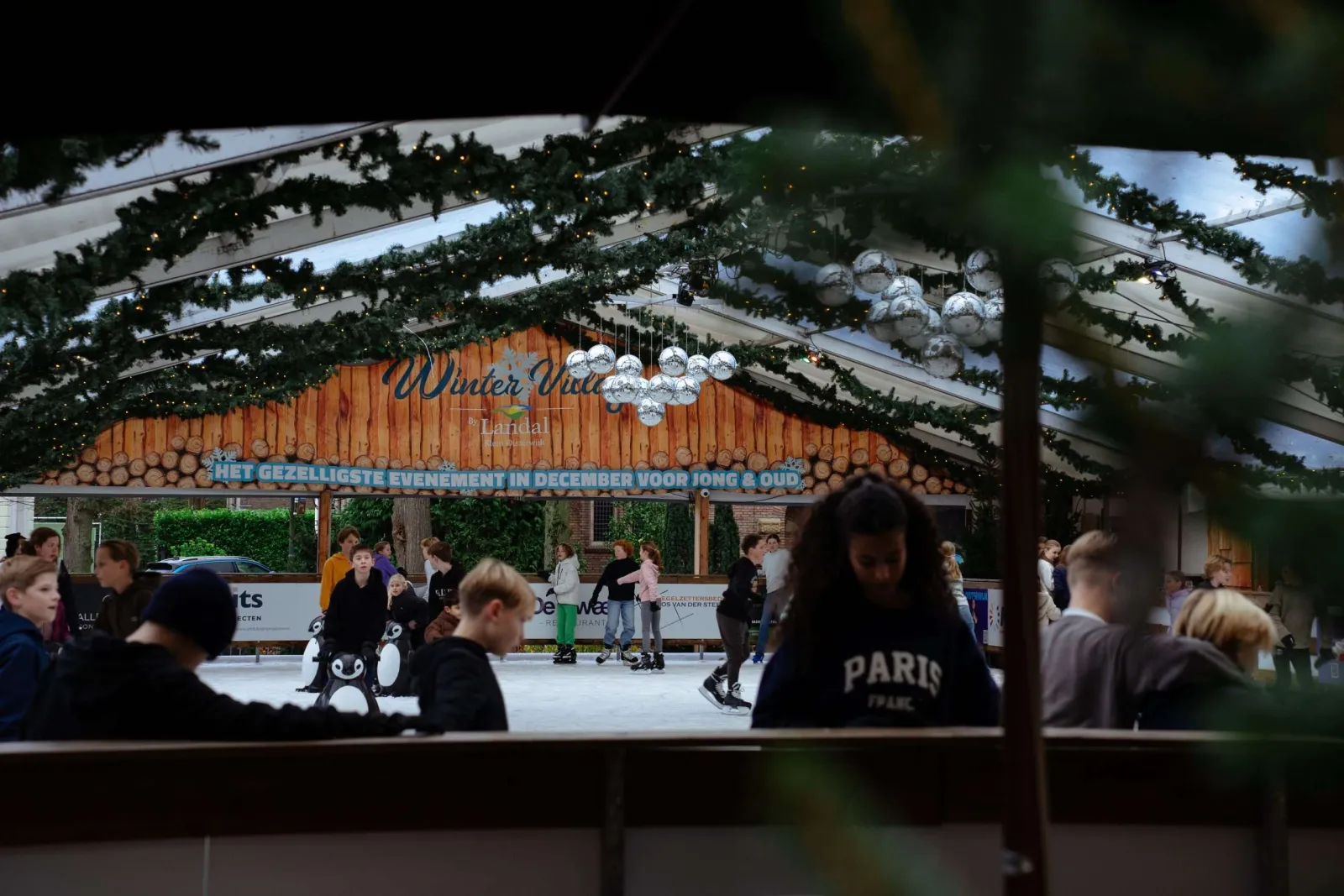 Skaters on the covered Winter Village Oisterwijk rink, seen through the Christmas decorations toward the wooden Winter Village entrance sign; people at distance, not posing. — Winter Village Oisterwijk