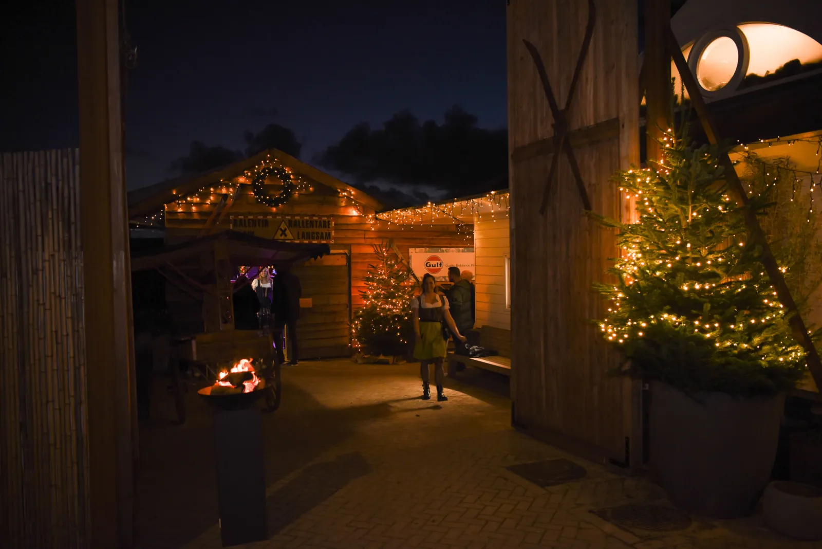 Evening view into the Winter Wonderland yard at Beachclub C, with lit fir trees and timber huts around a brazier. — Winter Wonderland Noordwijk
