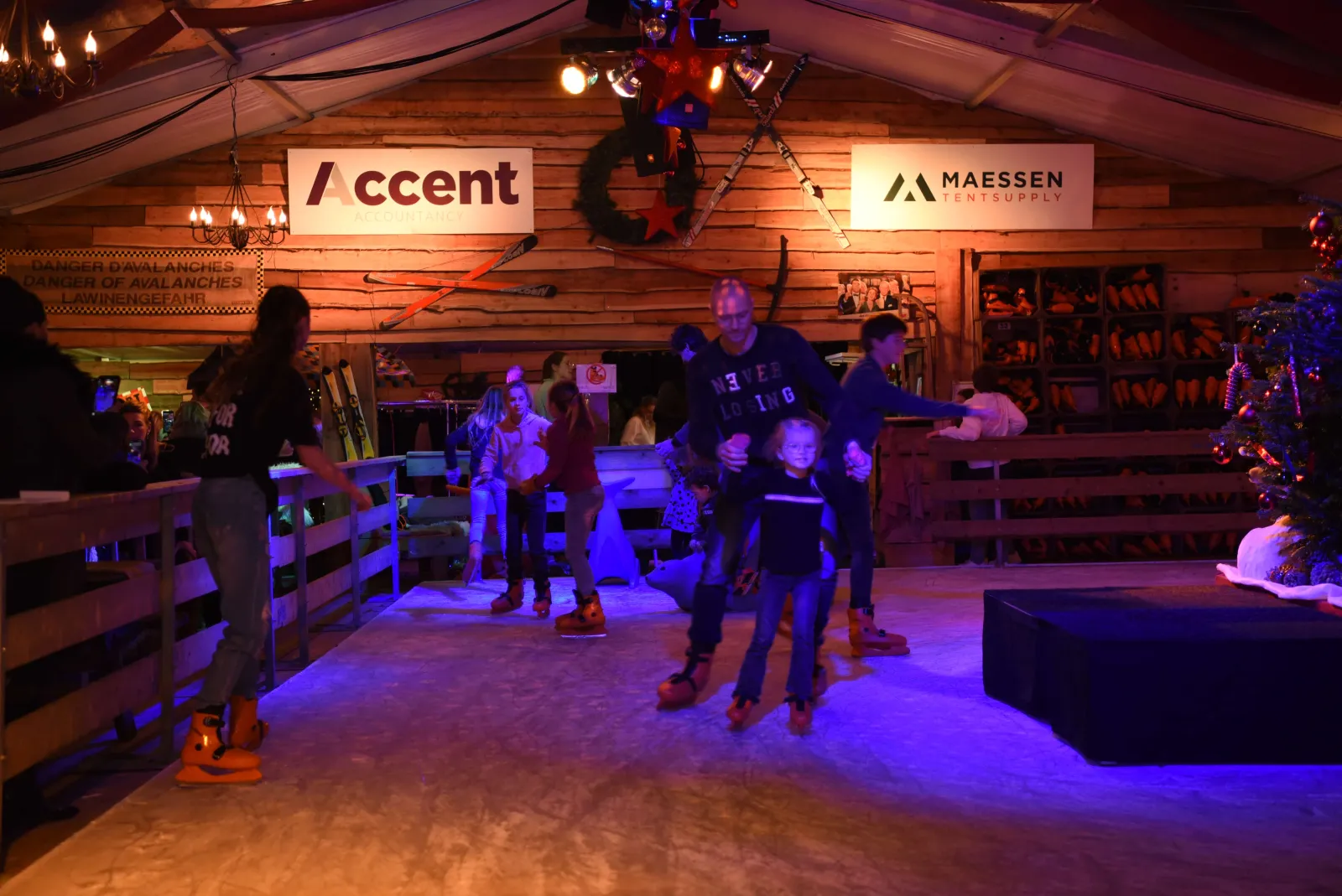 Skaters on the 120 m2 real-ice rink inside the heated Alpenstube at Beachclub C, under timber walls and stage lighting. — Winter Wonderland Noordwijk