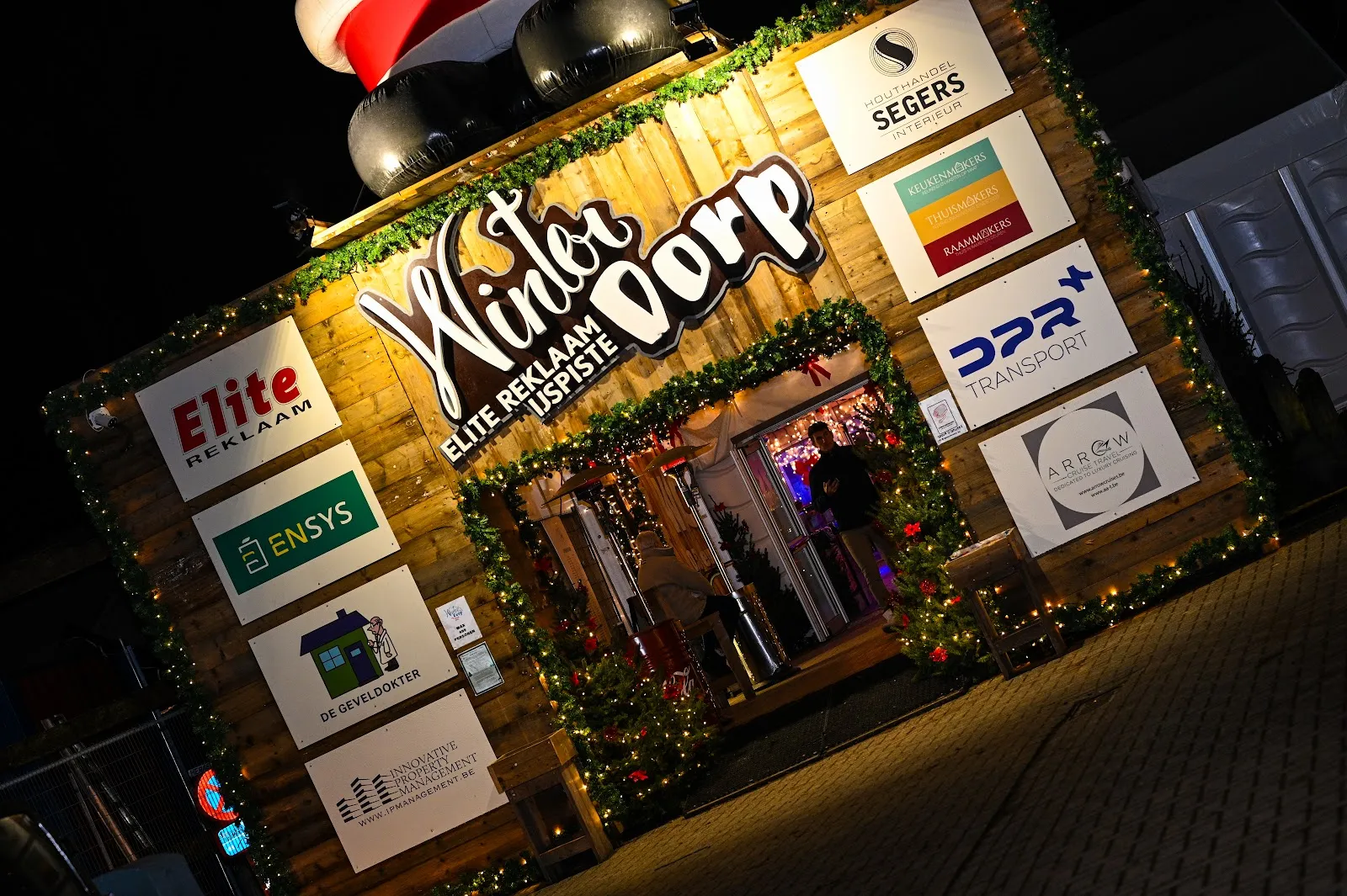 The wooden WinterDorp gate in Schilde at night with the sponsor boards and the lit entrance – Winterdorp Schilde