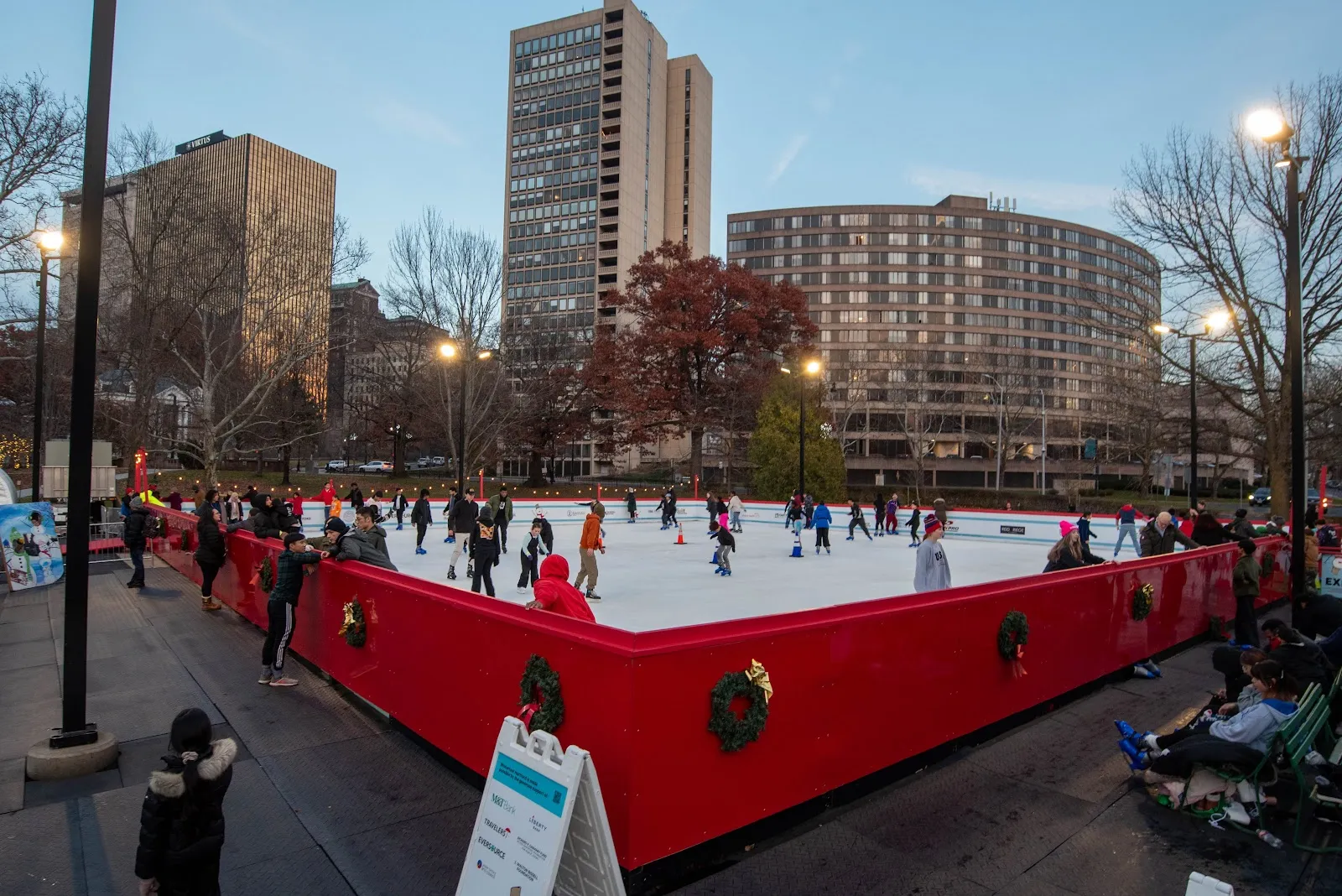 The rink at dusk seen from the walkway, its red boards hung with wreaths, with skaters on the ice and the apartment towers and street lamps beyond. — Winterfest Hartford