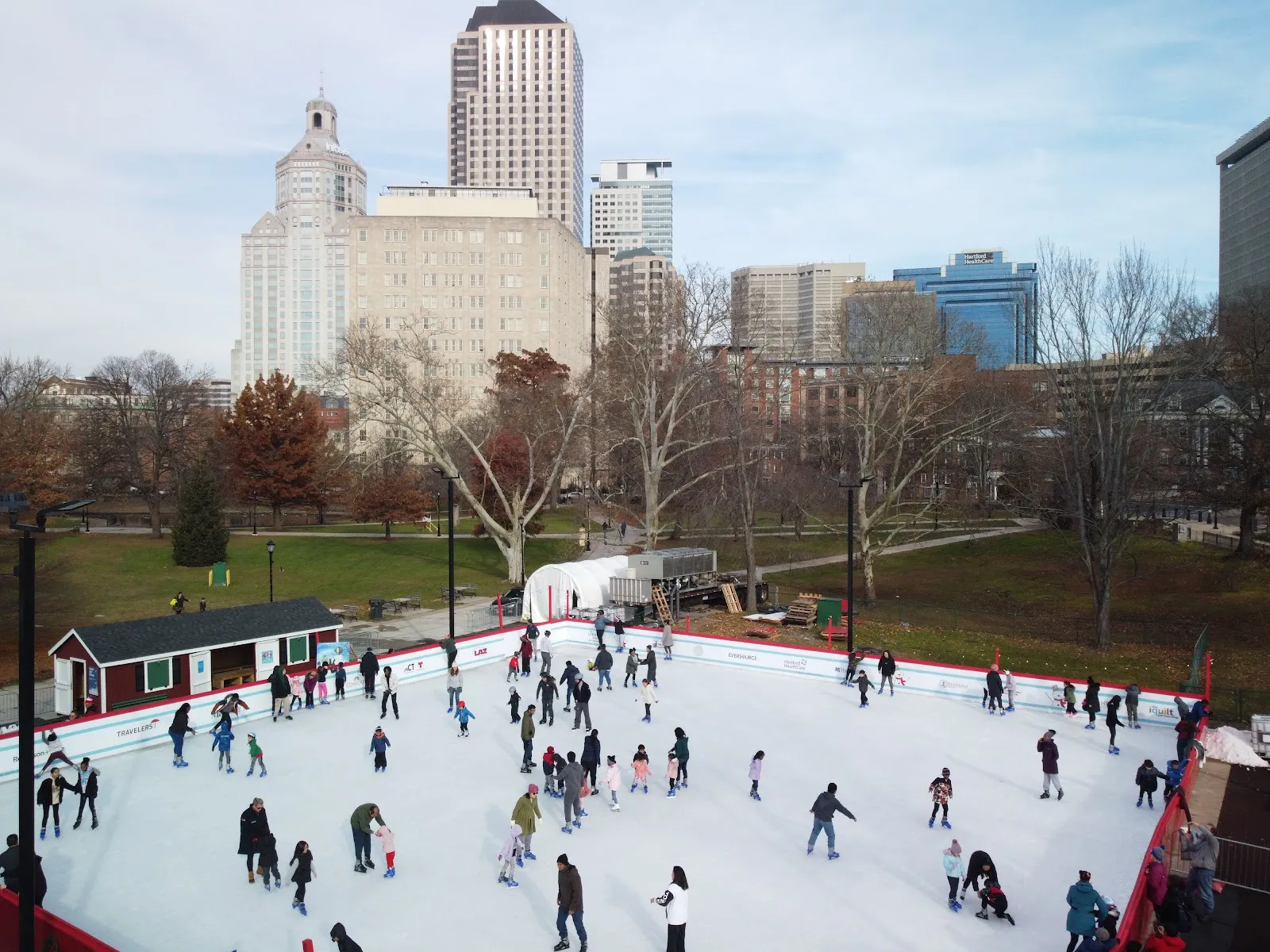 The square rink from above, full of skaters, with the skate shack at the left and Hartford's downtown buildings behind the bare trees of Bushnell Park. — Winterfest Hartford