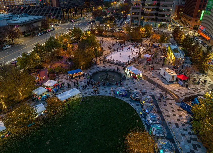 Una vista aérea de Kiener Plaza en otoño con la pista de patinaje, las casetas de comida y los asientos con domo dispuestos alrededor de la fuente central. — Winterfest Ice Rink at Kiener Plaza