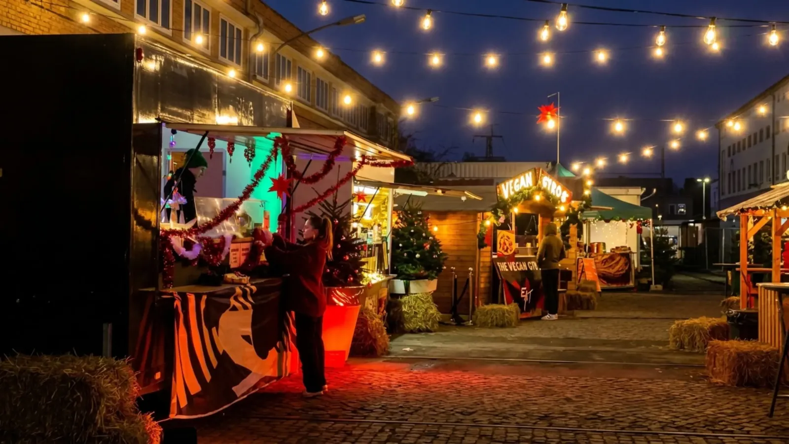 Die gepflasterte Marktgasse des Winterquartiers bei Nacht, mit Holzbuden und Lichterketten neben der Eishalle. – Winterquartier Berlin Eisbahn