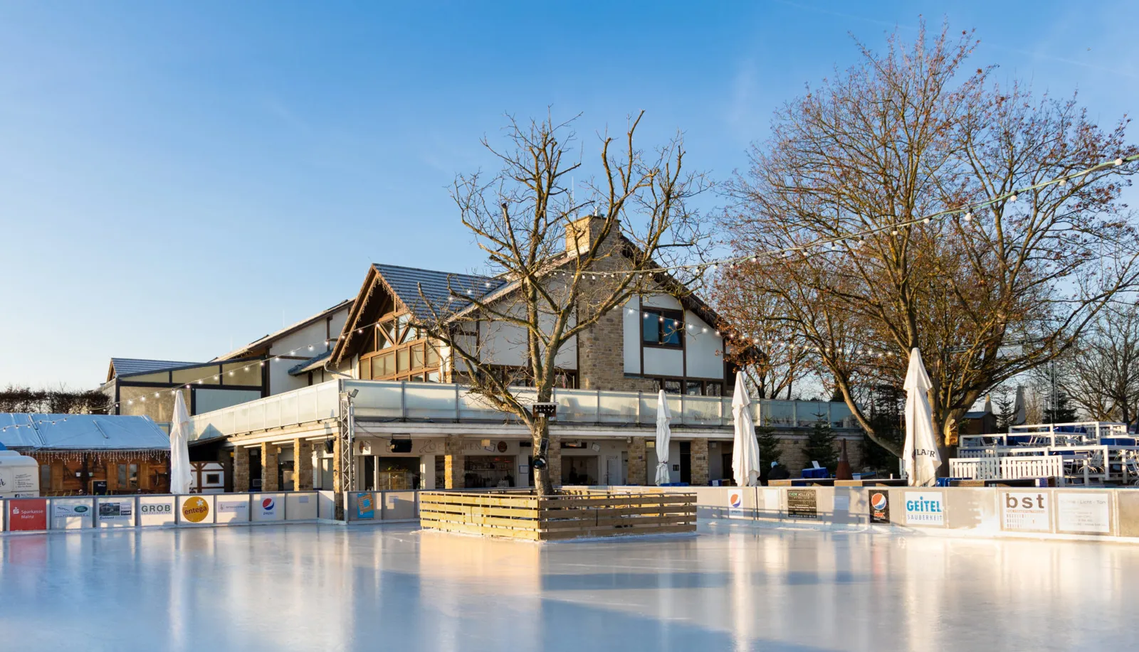 Das Fachwerkgebäude des Hofguts hinter der Eisbahn an einem klaren Wintertag, mit kahlen Bäumen und geschlossenen Sonnenschirmen. – Winterwelt Panorama-Eisbahn Hofgut Laubenheimer Höhe