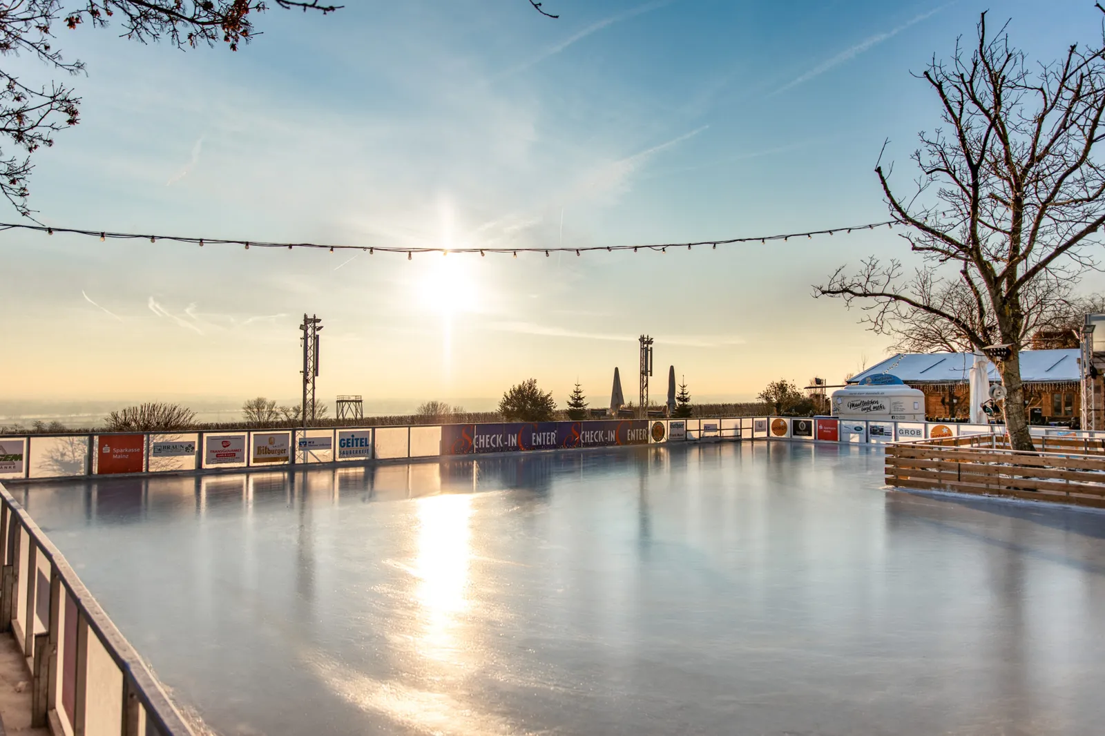 Die leere Freiluft-Eisfläche bei Sonnenuntergang, Banden mit Werbetafeln, eine Lichterkette und kahle Bäume. – Winterwelt Panorama-Eisbahn Hofgut Laubenheimer Höhe