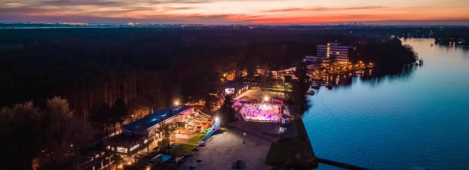 Das Strandbad Grünau am Wasser bei Einbruch der Nacht, mit der beleuchteten Eisbahn der Winterwelt und den Gebäuden des Bades zwischen den Bäumen. – Winterwelt Grünau (Strandbad Grünau)