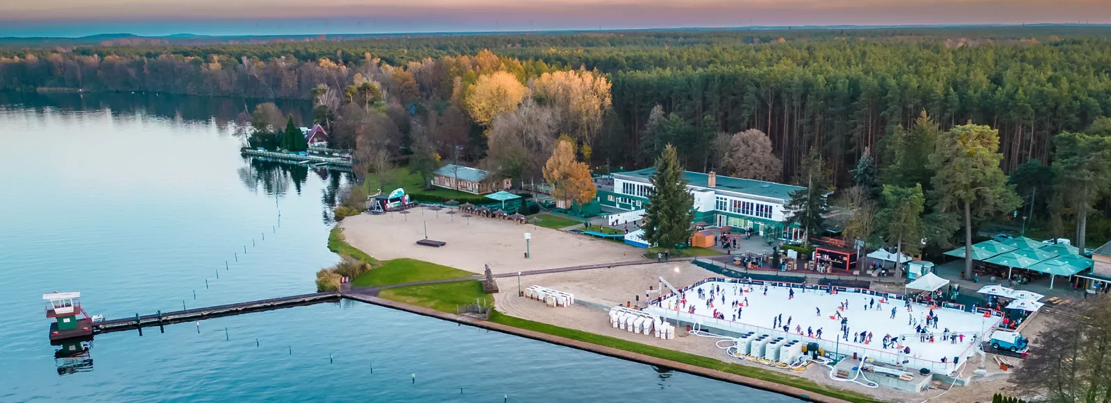 Luftaufnahme der Eisbahn der Winterwelt Grünau am Ufer der Dahme in der Dämmerung, mit Schlittschuhläufern auf dem 1.000 m² großen Eis neben dem Strand und den Gebäuden des Bades. – Winterwelt Grünau (Strandbad Grünau)