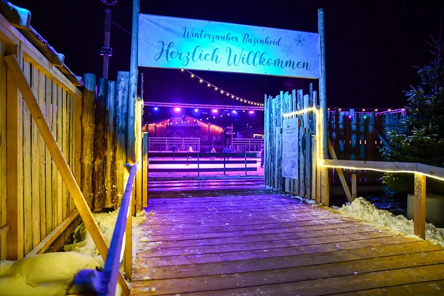 The wooden entrance gate of the Winterzauber Bazenheid winter village with its 'Herzlich Willkommen' banner at night; no people. — Winterzauber Bazenheid