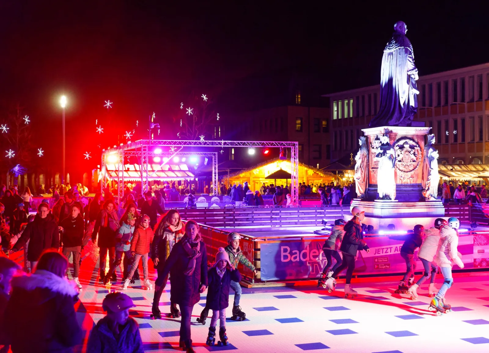Die Eisbahn der Winterzeit auf dem Karlsruher Friedrichsplatz bei Nacht, Schlittschuhläufer auf dem beleuchteten Eis um die Statue, dahinter Marktstände und Flutlicht. – Stadtwerke Karlsruhe WINTERZEIT