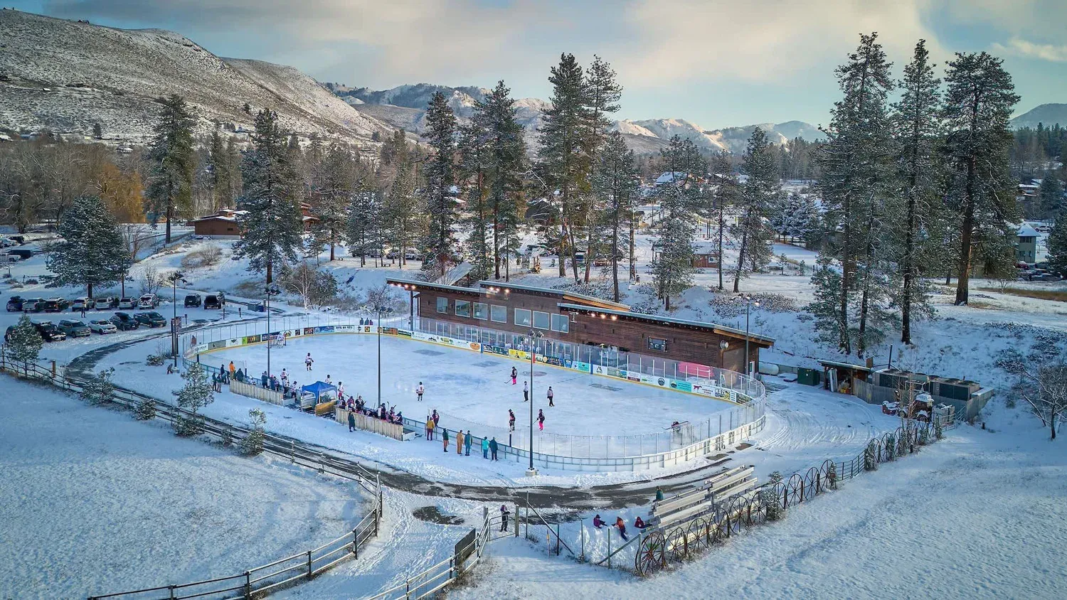 Aerial view of the Winthrop Rink in snow, the outdoor ice and its clubhouse building set among pine trees below the mountains — Winthrop Rink