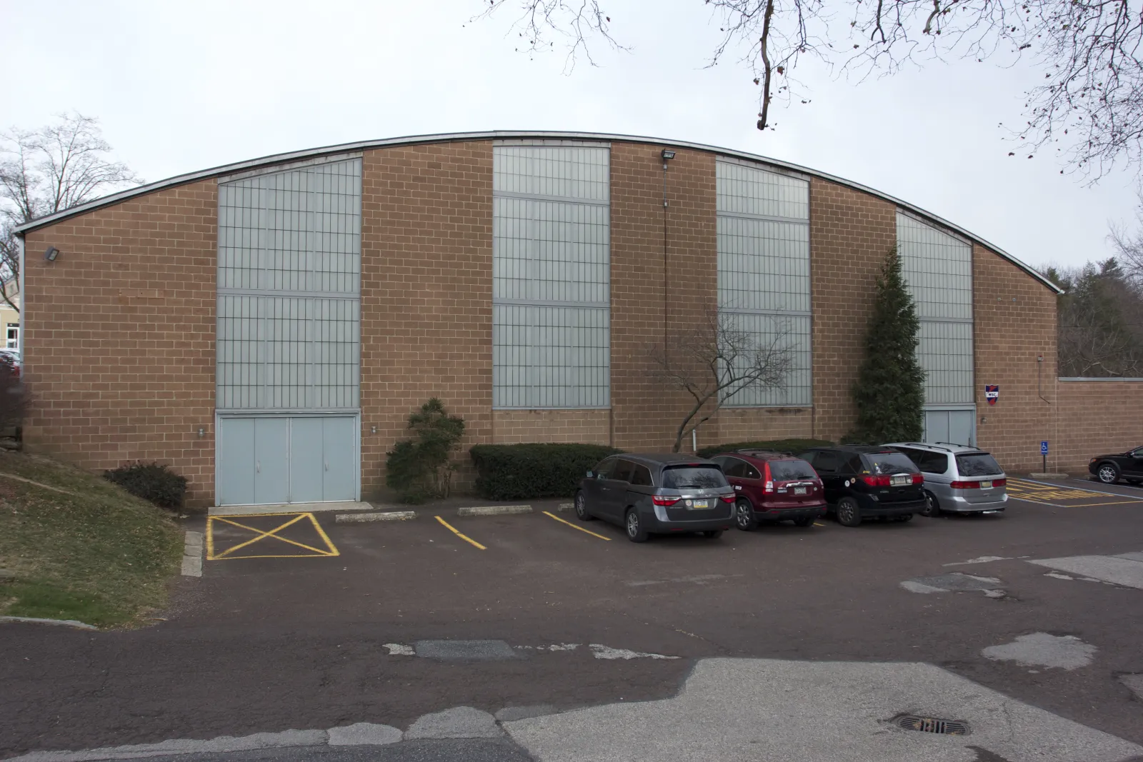 The 1950s brick rink building with its curved roofline and tall glass-block window panels, cars parked along the side. — Wissahickon Skating Club