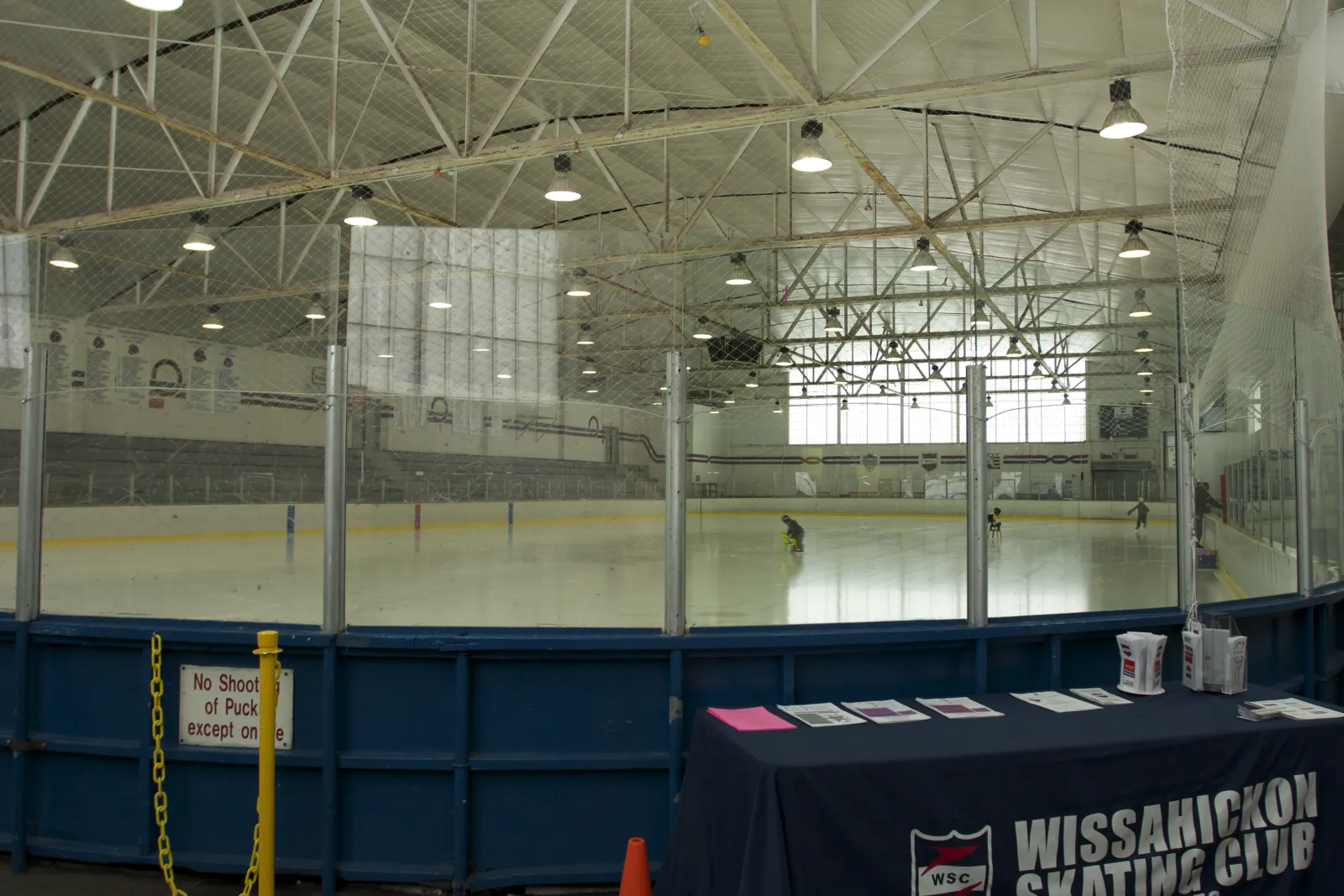 The rink under its white steel roof trusses, seen over the blue dasher boards, with a Wissahickon Skating Club table in the foreground. — Wissahickon Skating Club