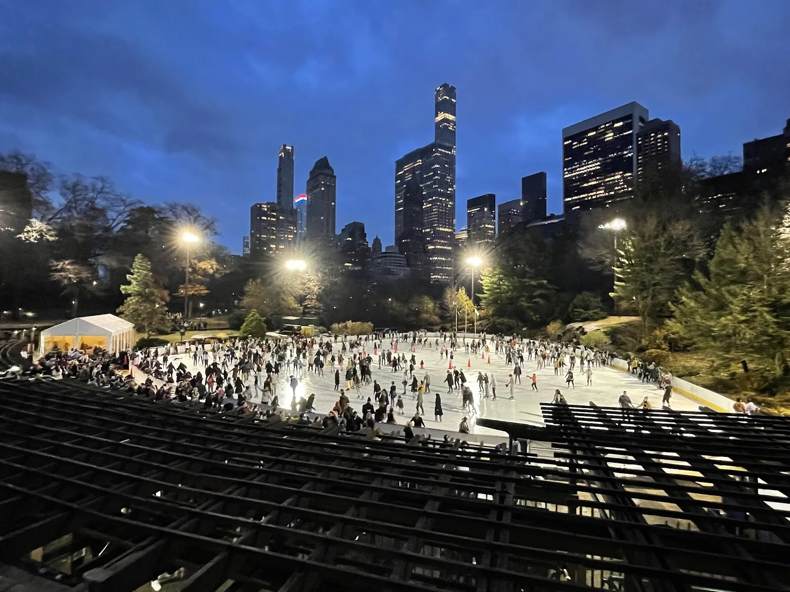 Wollman Rink de noche, con patinadores repartidos por el hielo iluminado debajo de las gradas escalonadas y el horizonte de Midtown brillando detrás de los árboles. — Wollman Rink