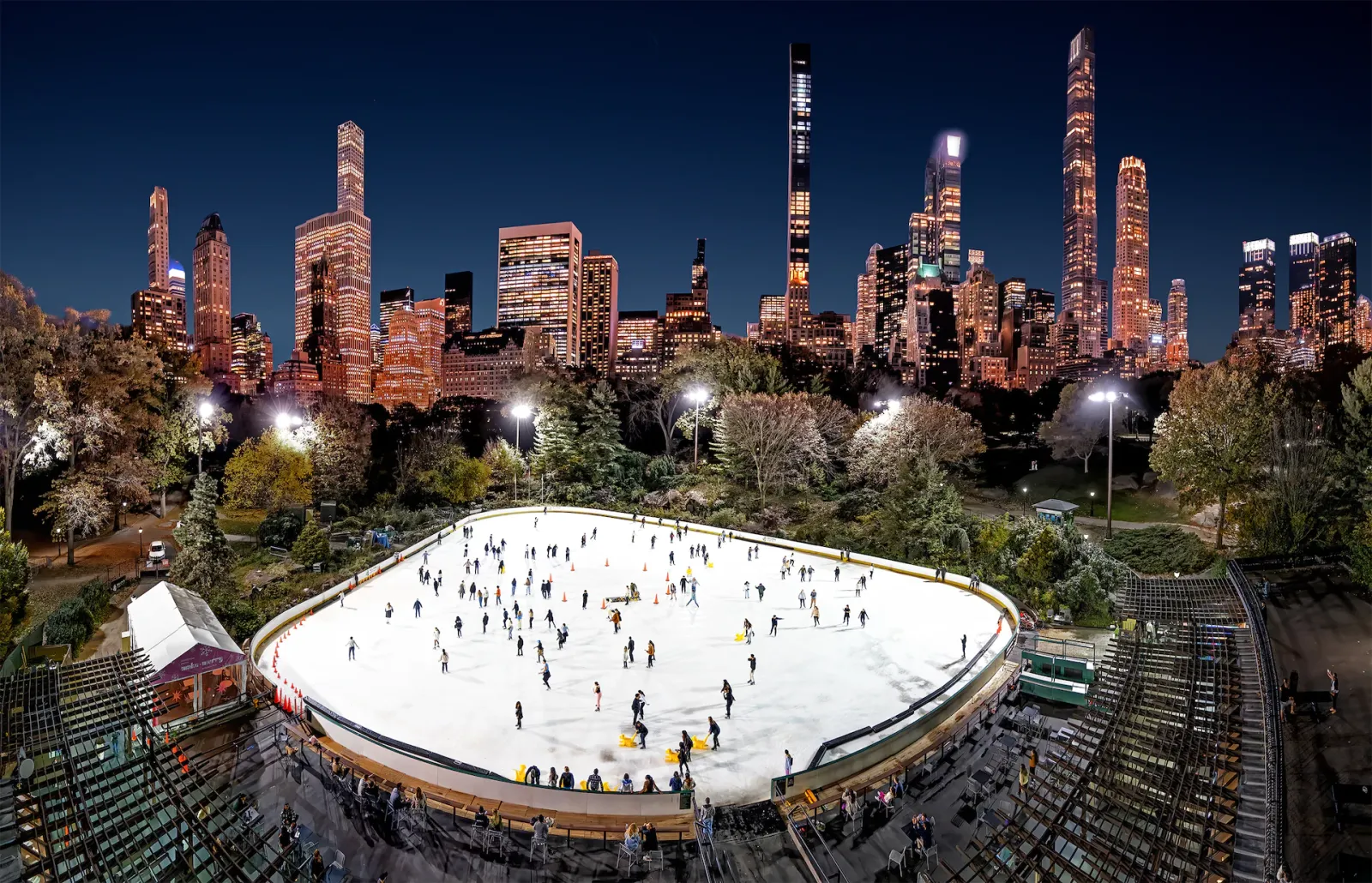 Una vista nocturna desde arriba de toda la pista ovalada llena de patinadores, rodeada de árboles iluminados y de la tribuna, con el horizonte de Midtown y sus rascacielos altísimos al fondo. — Wollman Rink