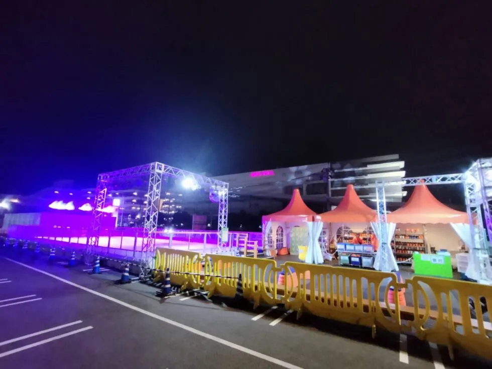夜のポップアップリンクの会場。駐車場の脇にオレンジ色のチケットテントとライトの入ったリンクの囲いが並びます。 — Wonder Rink AEON Mall Kashihara