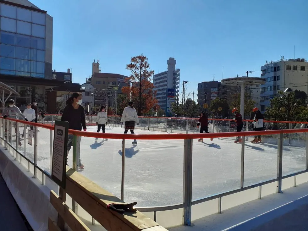 The outdoor rink on a sunny afternoon with distant skaters, city buildings behind and a railing in the foreground. — Wonder Rink Ario Kameari