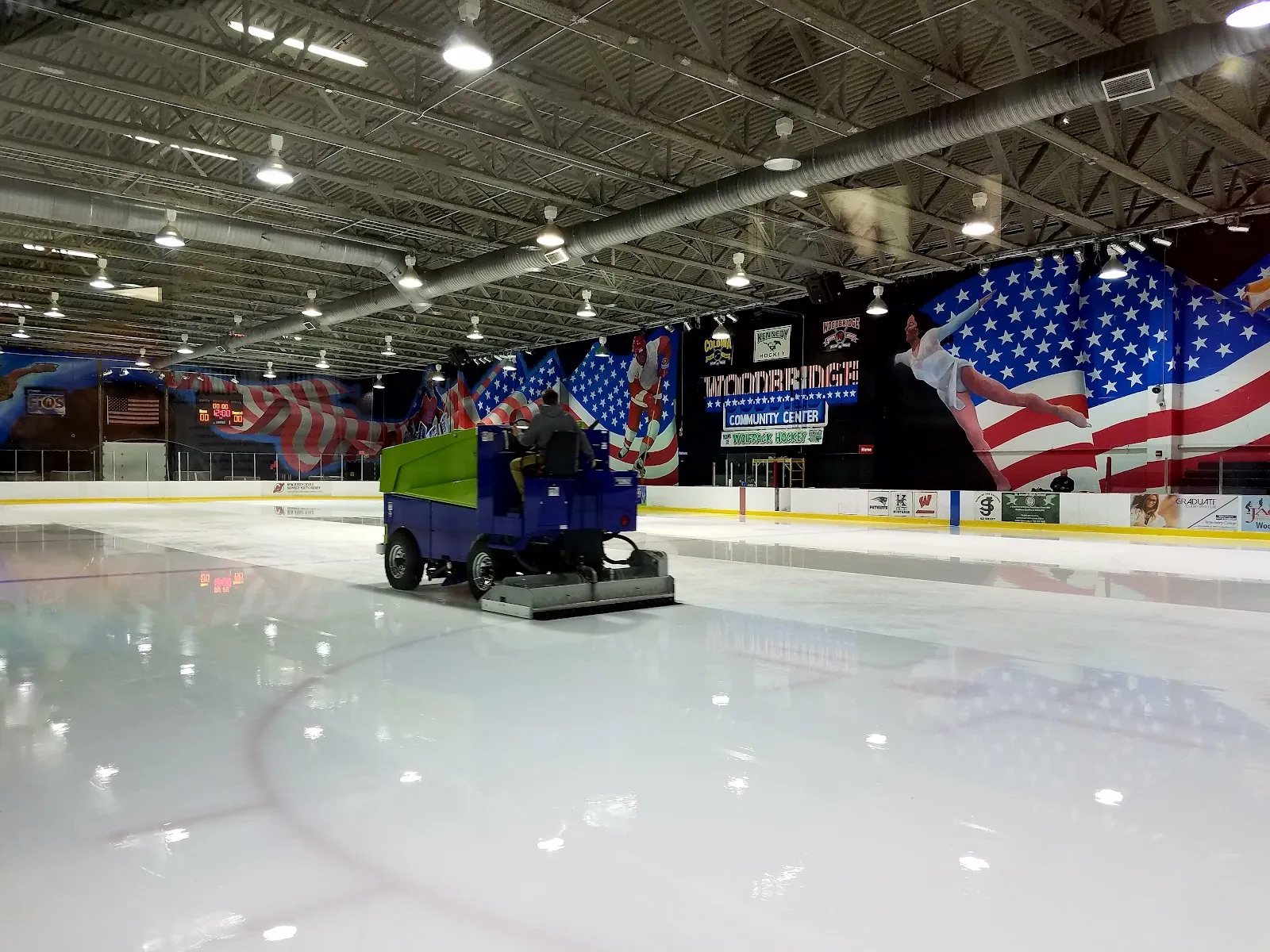 An ice resurfacer crossing the freshly flooded rink, with a large American flag mural and a Woodbridge Community Center banner along the far wall. — Woodbridge Community Center