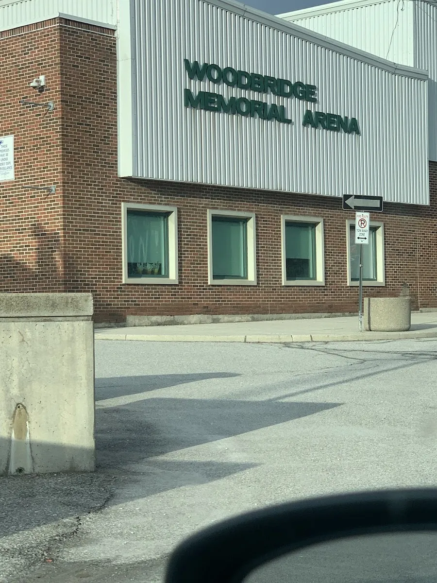 La esquina del edificio del Woodbridge Memorial Arena con su nombre en letras verdes sobre el revestimiento metálico, encima de un ala de ladrillo con tres ventanas, vista desde el aparcamiento. — Woodbridge Pool & Memorial Arena (Vaughan)