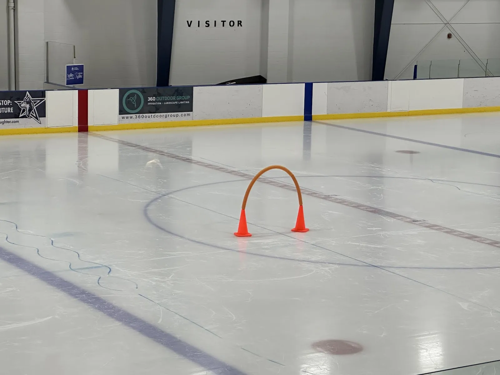 El hielo vacío del Woodbridge Pool and Memorial Arena, con un arco de entrenamiento naranja sobre la pista y el letrero de Visitor sobre las vallas. — Woodbridge Pool & Memorial Arena (Vaughan)