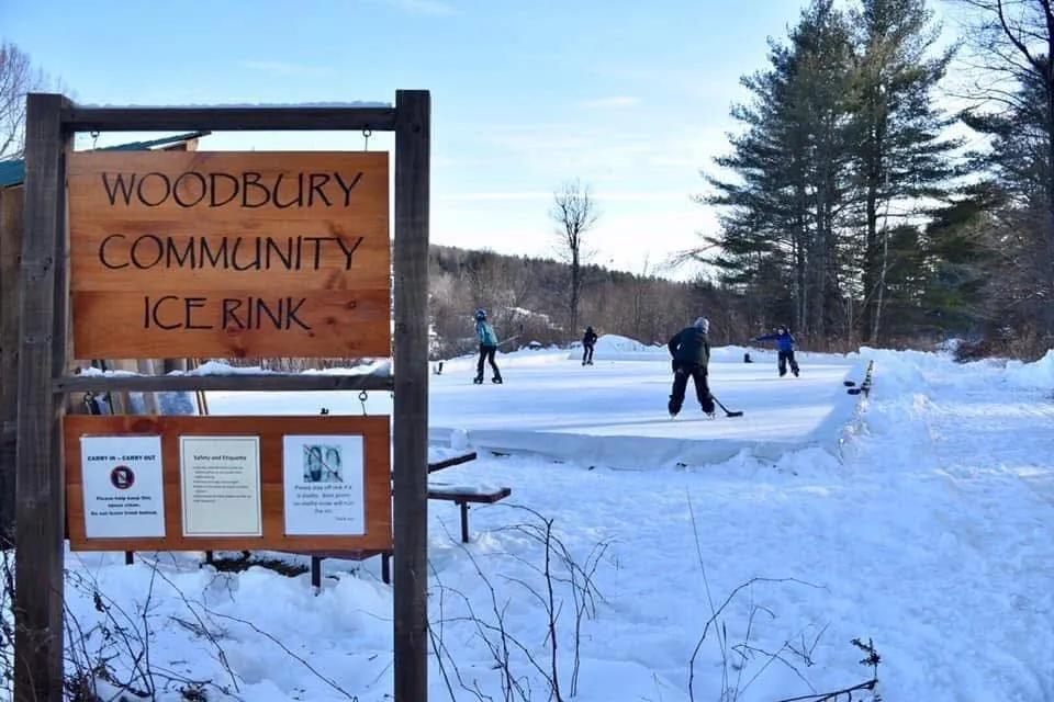La pista comunitaria Woodbury Community Ice Rink en Vermont: el letrero de la pista a la izquierda y unos pocos patinadores en el hielo nevado a lo lejos. — Woodbury Community Ice Rink