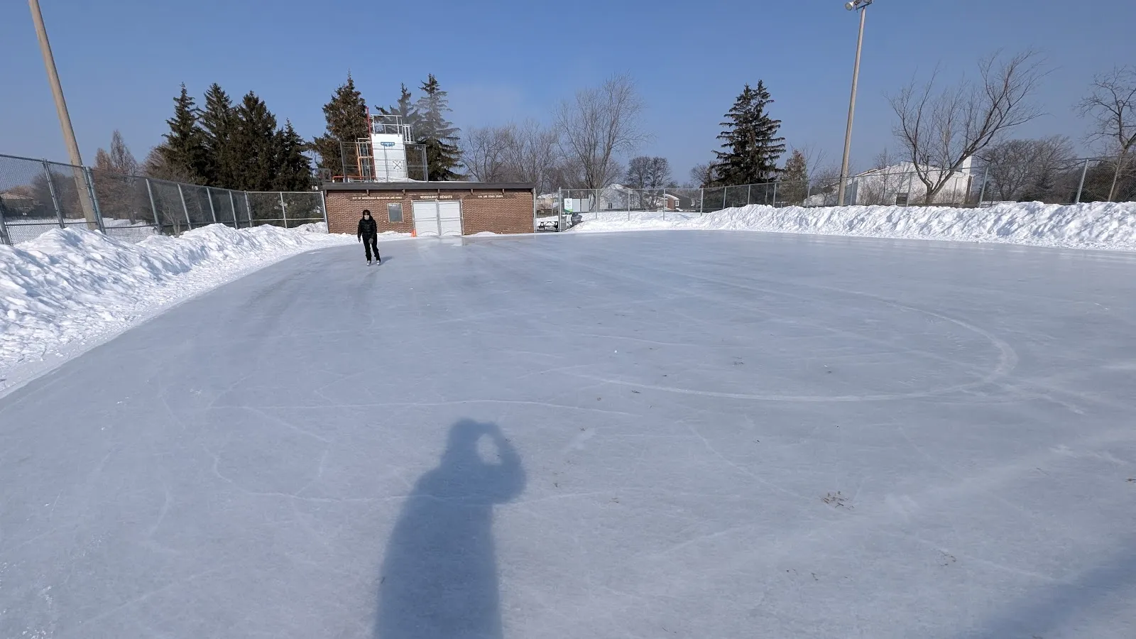 The Woodhurst Heights Park outdoor rink ice on a sunny day with snowbanks and the park building behind, one distant skater — Woodhurst Heights Outdoor Rink